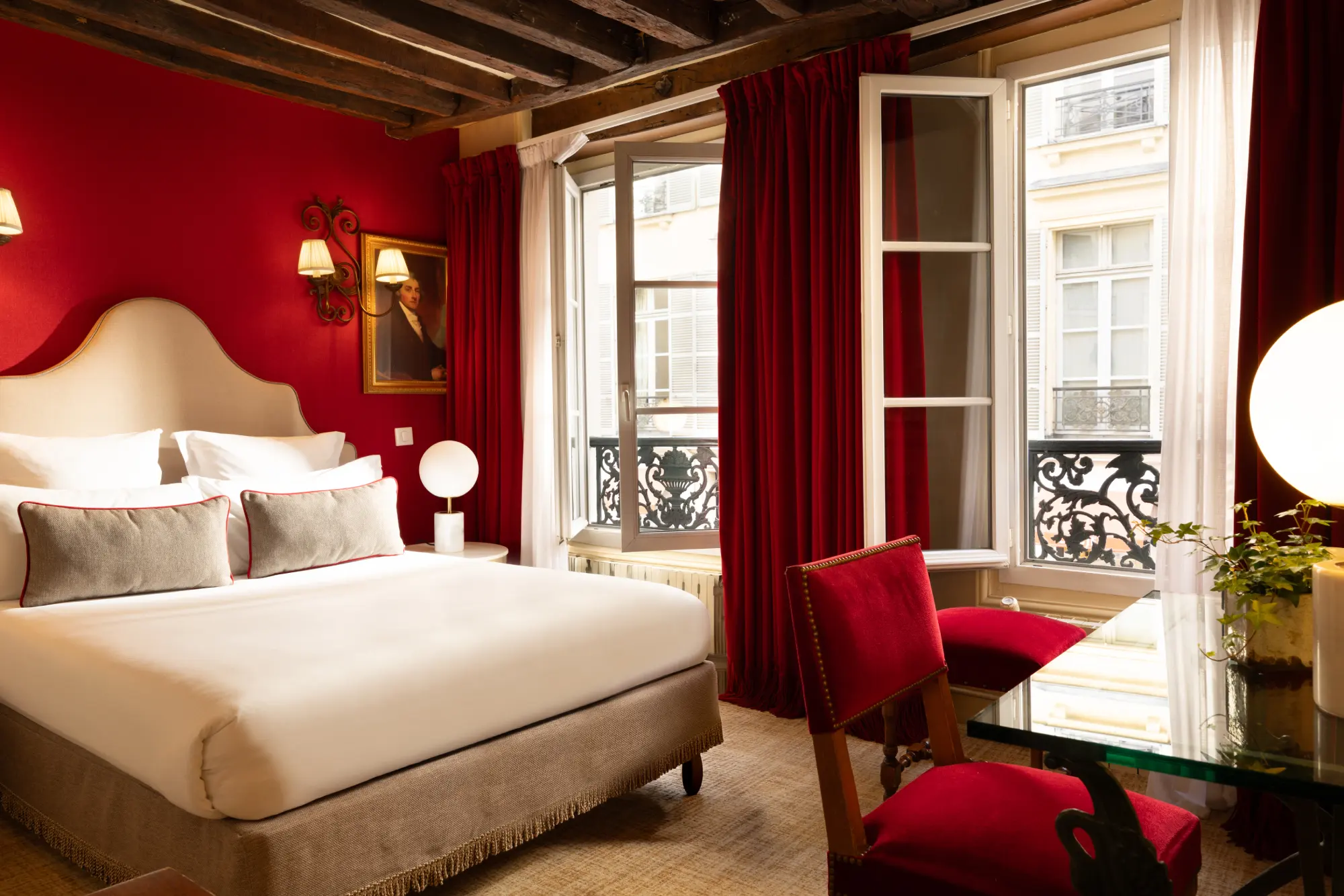 Chambre au mur rouge avec fenêtres ouvertes sur un balcon en fer forgé donnant sur une rue parisienne
