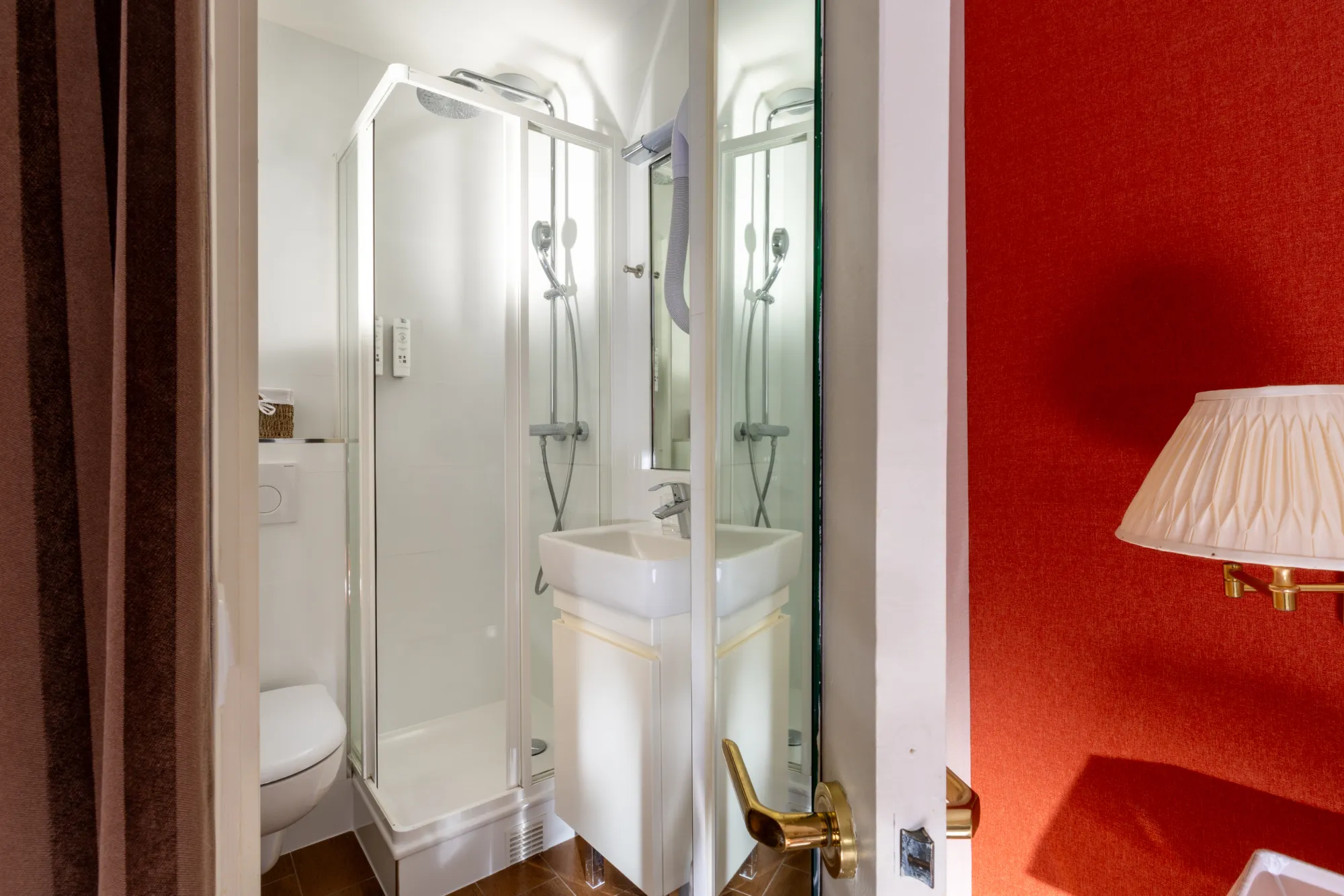 Douche vitrée et vasque blanche dans la salle d'eau de la chambre Cosy, avec vue sur le mur rouge de la chambre à l'Hôtel des Deux Continents