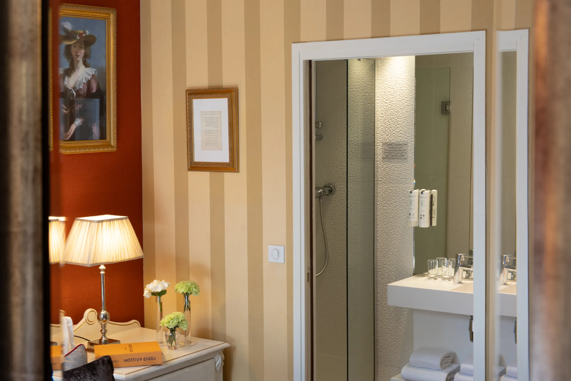Salle de bain à rayures avec douche vitrée, tableau ancien et coiffeuse fleurie dans la chambre familiale