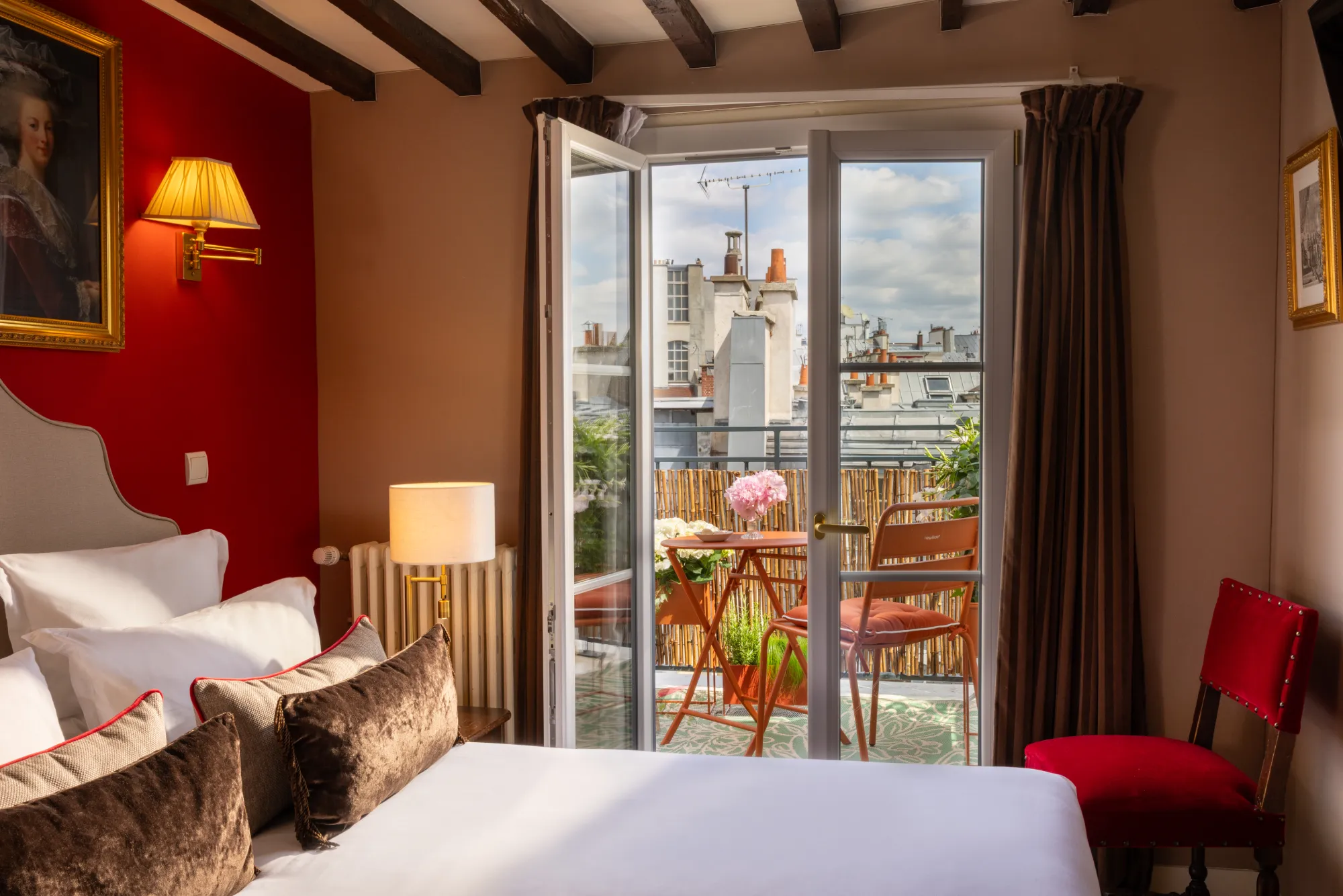 Chambre au mur rouge et poutres apparentes avec portes-fenêtres ouvertes sur une terrasse fleurie, Hôtel des Deux Continents