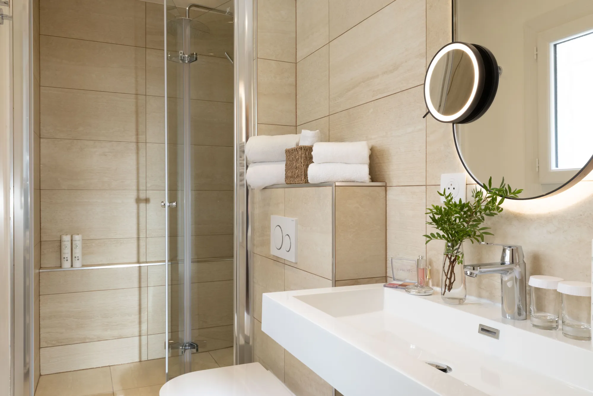 Douche à l'italienne vitrée dans une salle de bain en pierre beige avec vasque et miroir rond, Hôtel des Deux Continents